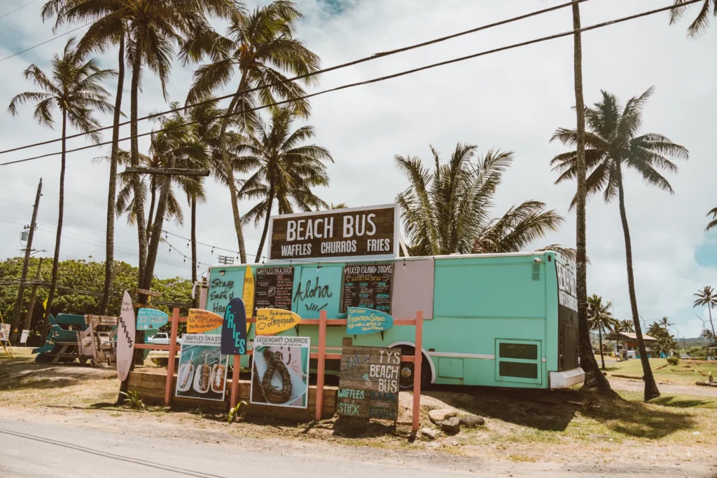 Beach bus food truck in Hawaii offering waffles, churros, and fries under palm trees.