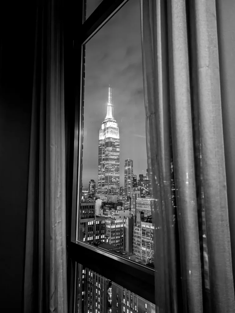 Black and white image of the Empire State Building seen through a window with curtains at night.