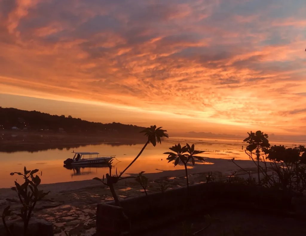 Breathtaking sunset in Nusa Penida, Bali with a scenic view of the bay and silhouetted palm trees.