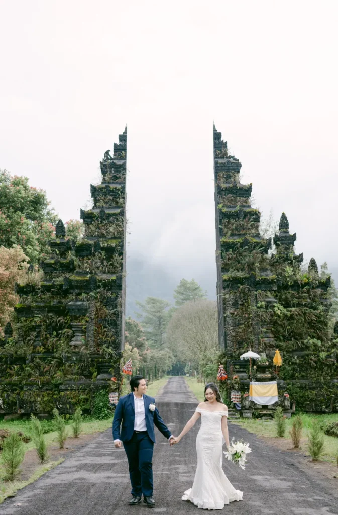 Bride and groom walking at traditional Balinese temple gates in a scenic landscape.