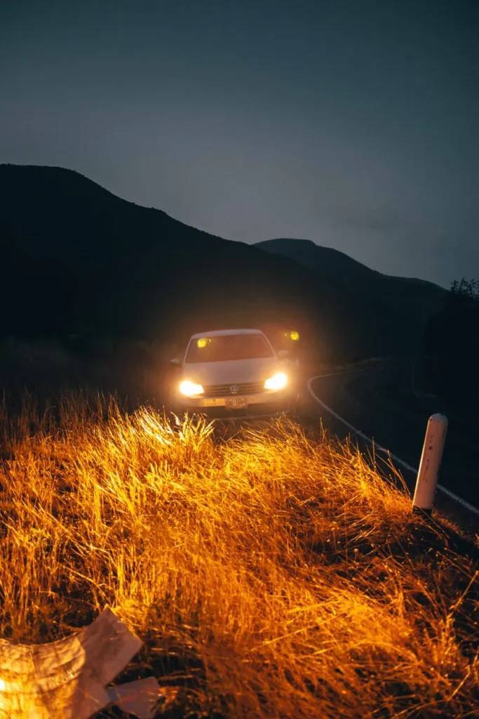 Car with headlights on, shining light over grassy roadside at night.