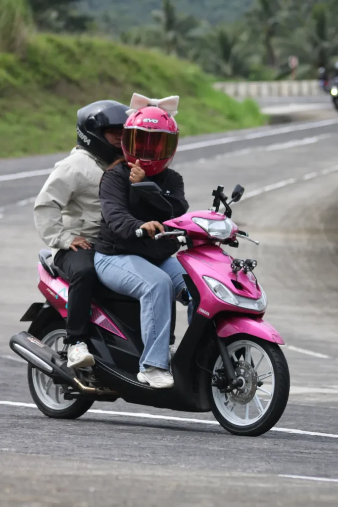 Couple enjoying a ride on a pink scooter along a scenic road. Wear helmets for safety.