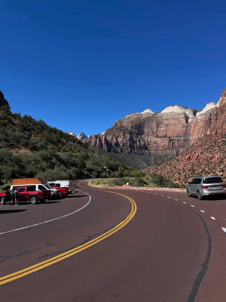 Curved road with parked cars and Zion Canyon mountains in background.