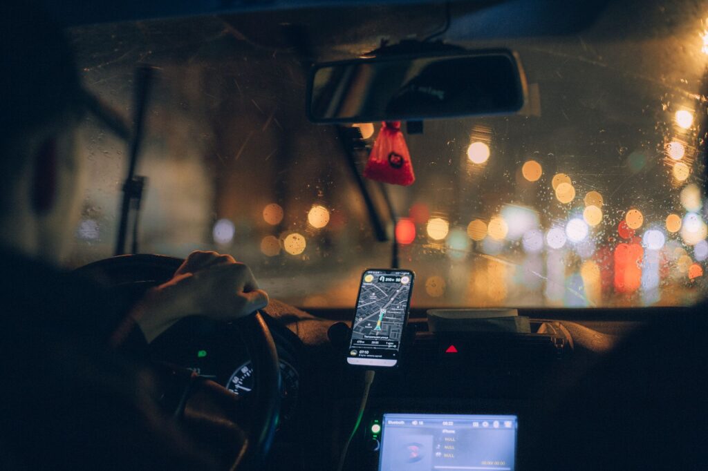 Driving at night in a car with GPS navigation on smartphone, city lights reflecting on wet windshield.