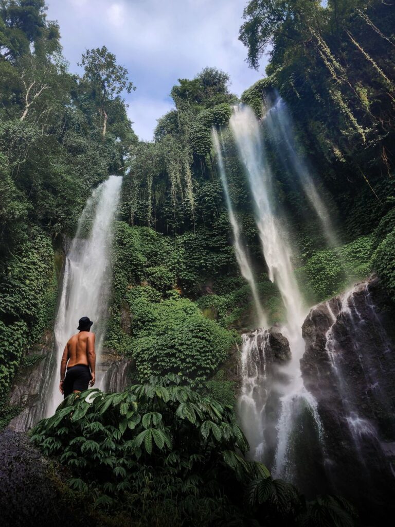 Explorer standing before a vibrant waterfall in Bali's lush rainforest.
