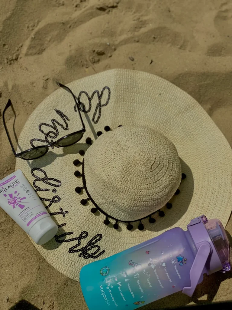 Flat lay of beach essentials: straw sun hat, sunglasses, sunscreen, and water bottle on sand.