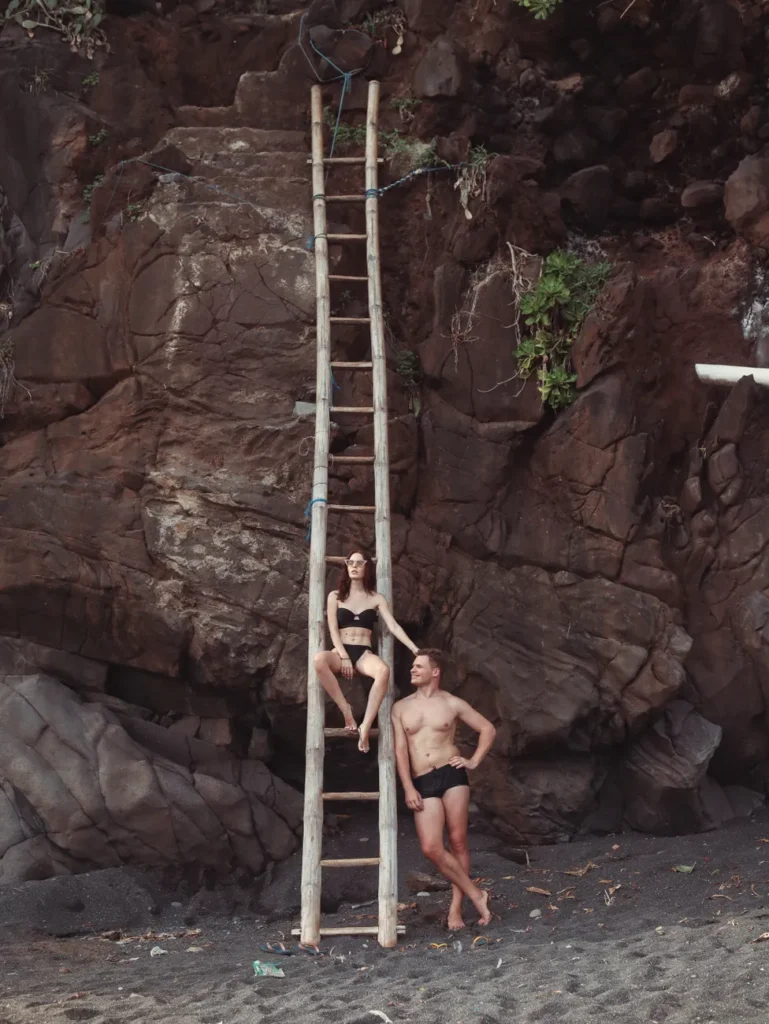 Full length of relaxed young female in black swimwear sitting on bamboo ladder and confident man leaning on ladder while spending time in mountain beach