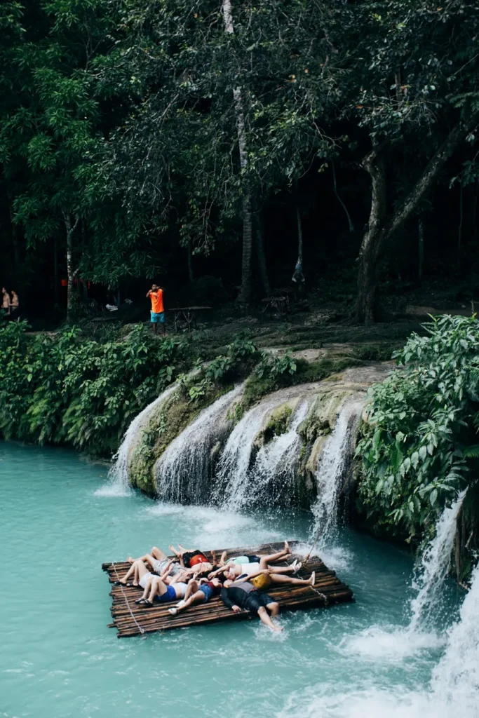 Group of people lounging on a bamboo raft near a scenic waterfall in a lush tropical setting.