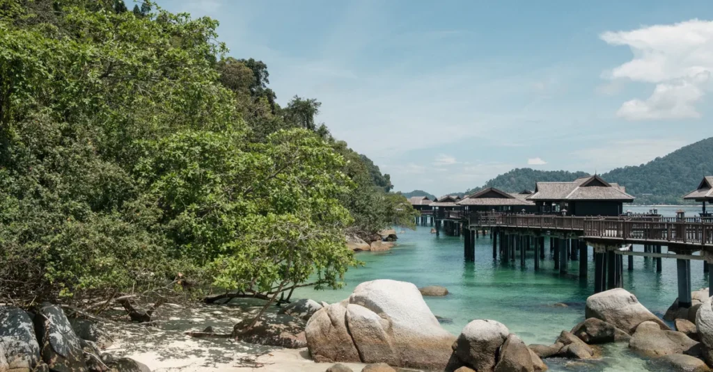 Ora Beach à deux (Seram, Maluku) : le guide concret pour snorkeler, dormir au calme et éviter les pièges Idyllic tropical beach with lush greenery, rocks, and overwater bungalows under clear skies.