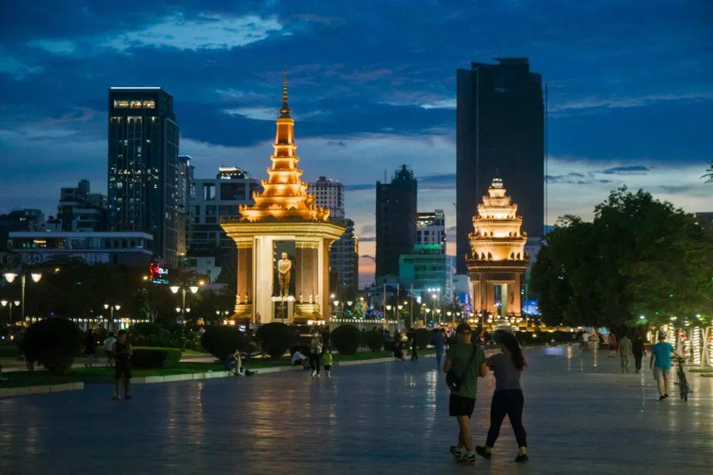 Illuminated Norodom Sihanouk Statue in Phnom Penh during night, with people walking nearby.