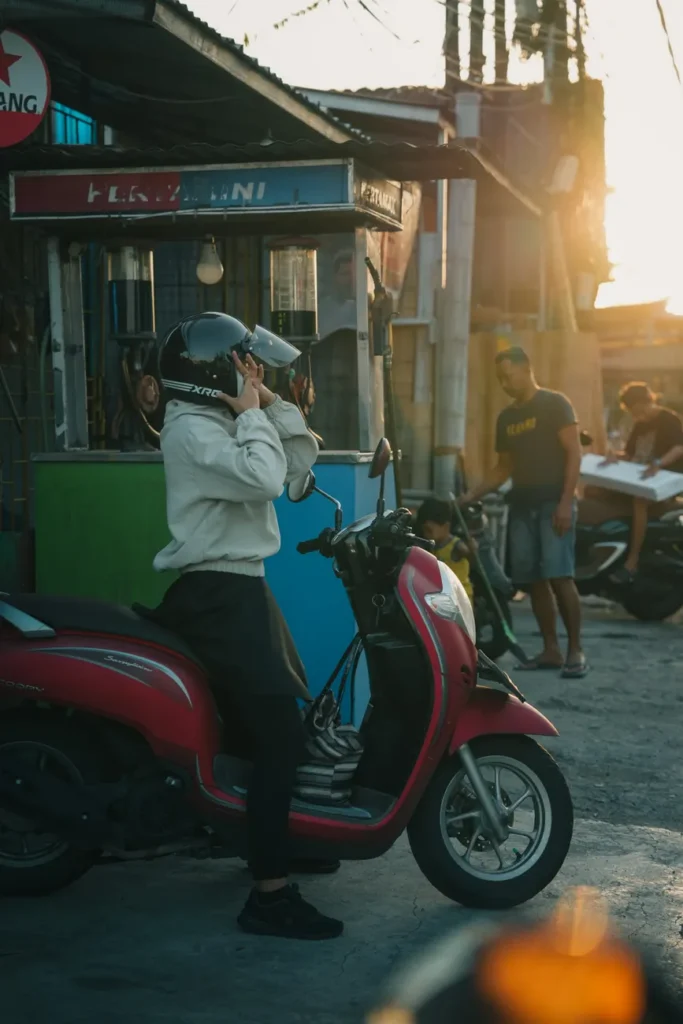 Man wearing helmet beside red scooter on a city street at sunset.