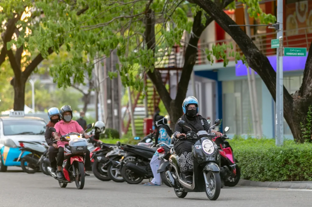 Motorcyclists navigating an urban street lined with trees on parked scooters.