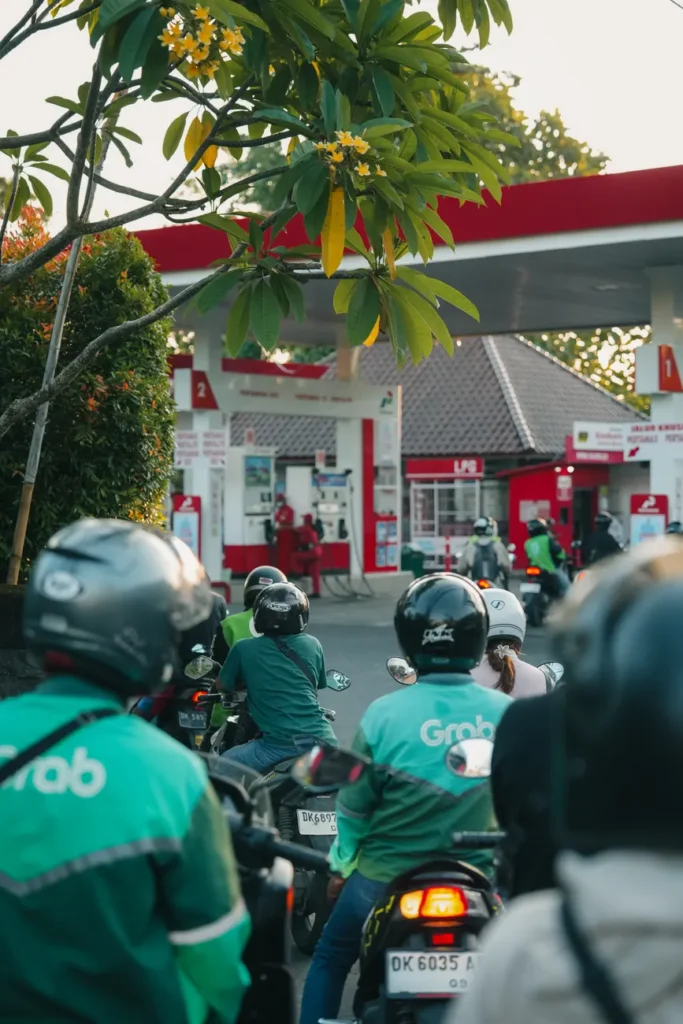 Motorcyclists wearing helmets lining up at a busy urban gas station under lush green trees.