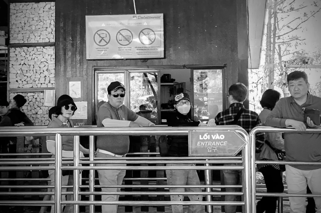 People waiting in line at an entrance with safety signage, captured in monochrome.