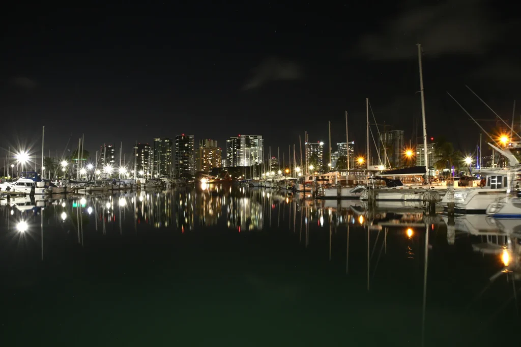 Reflective night view of Honolulu's marina with illuminated city skyline and boats along the harbor.