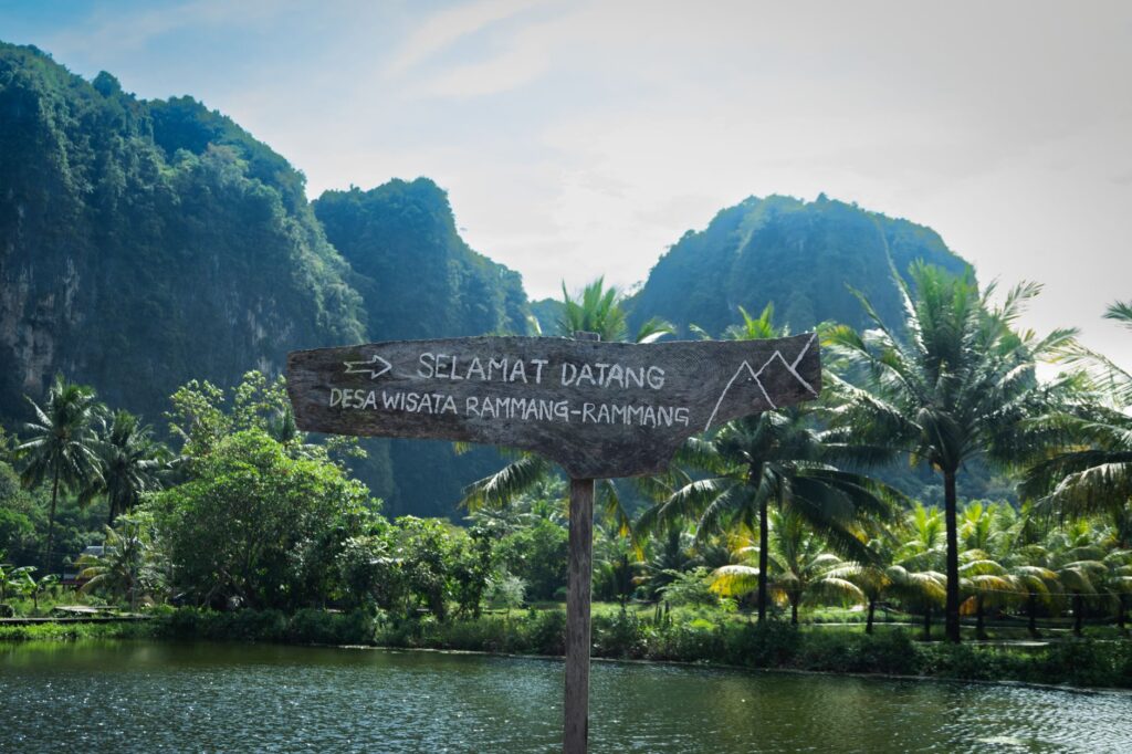Rustic sign welcoming visitors to scenic Rammang-Rammang village, surrounded by lush greenery and karst mountains.
