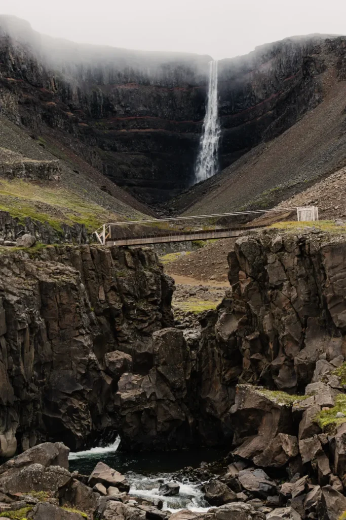 Scenic view of Hengifoss waterfall cascading down cliffs with a wooden footbridge crossing the rocky landscape.