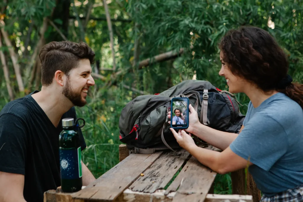 Side view of cheerful young female hiker in casual clothes sitting at wooden table in green forest and showing smiling boyfriend photos on smartphone while resting together during trekking