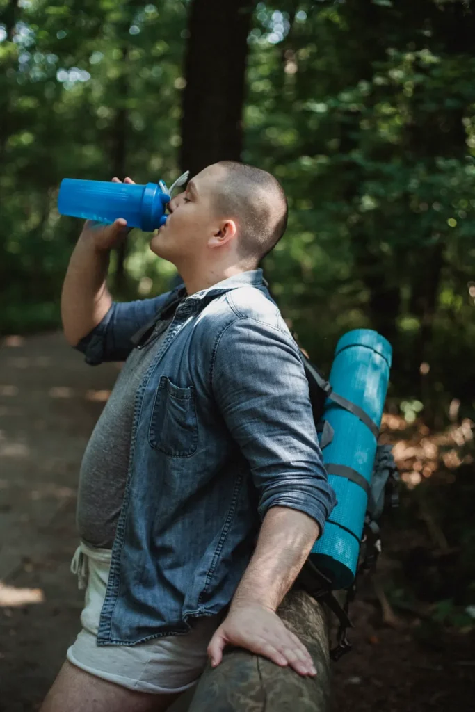 Side view of man in casual clothes with backpack drinking bottle of water while resting in park during hike in daytime