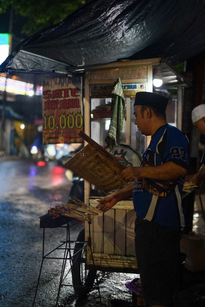 Street vendor grills satay at night market in rainy setting, creating an authentic street food experience.
