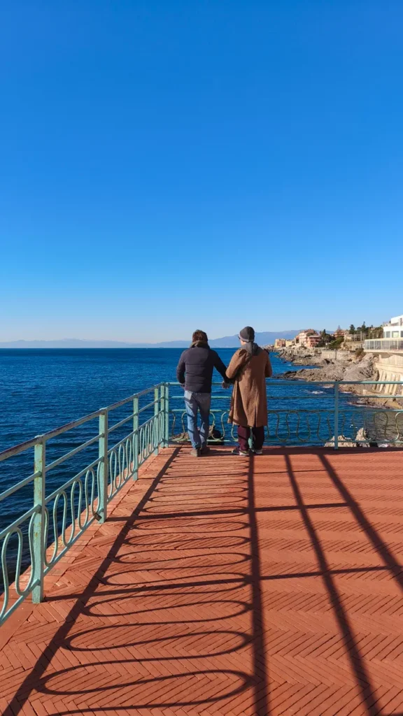Two adults walking hand in hand on a seaside promenade in Italy with a clear blue sky.