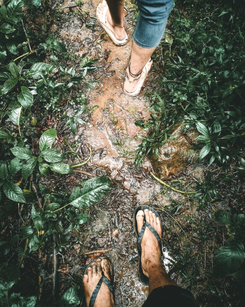 Two people walking on a muddy trail in a lush green forest, showcasing adventure and nature.