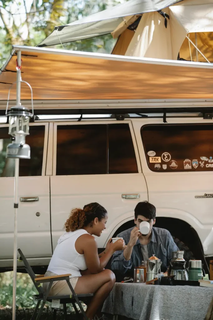Unrecognizable young multiethnic couple enjoying fresh coffee while sitting near SUV car with tent during camping in nature
