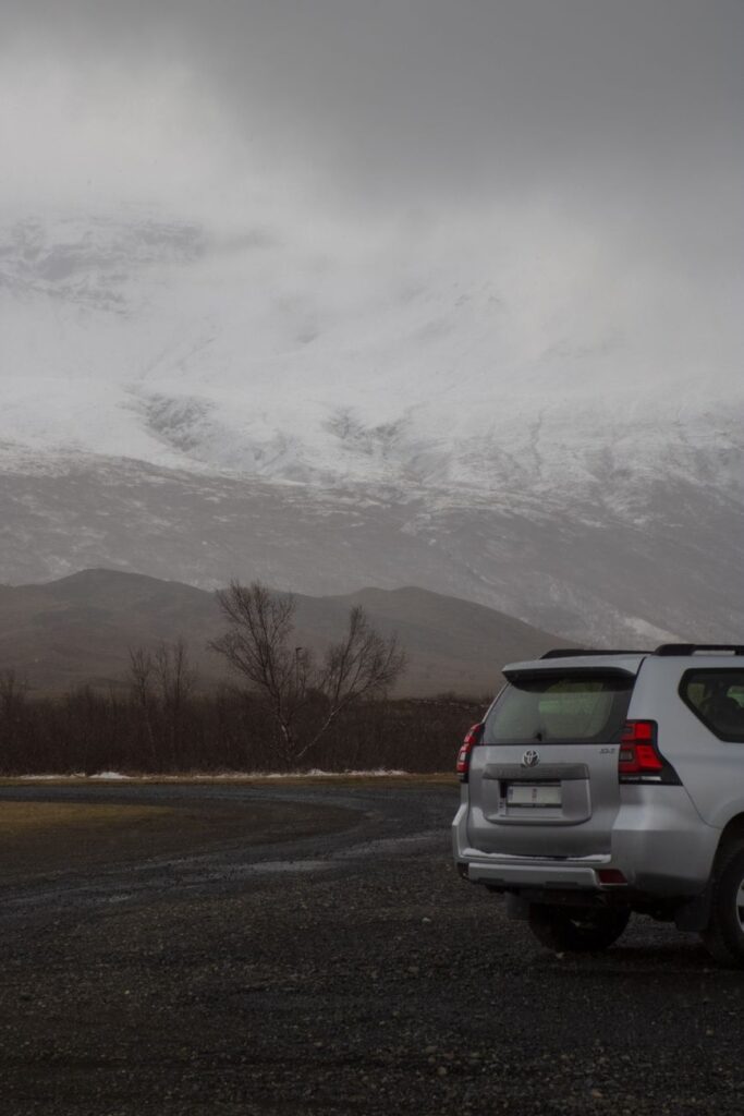 White off-road SUV parked on gravel path with snowy mountain backdrop under cloudy skies.