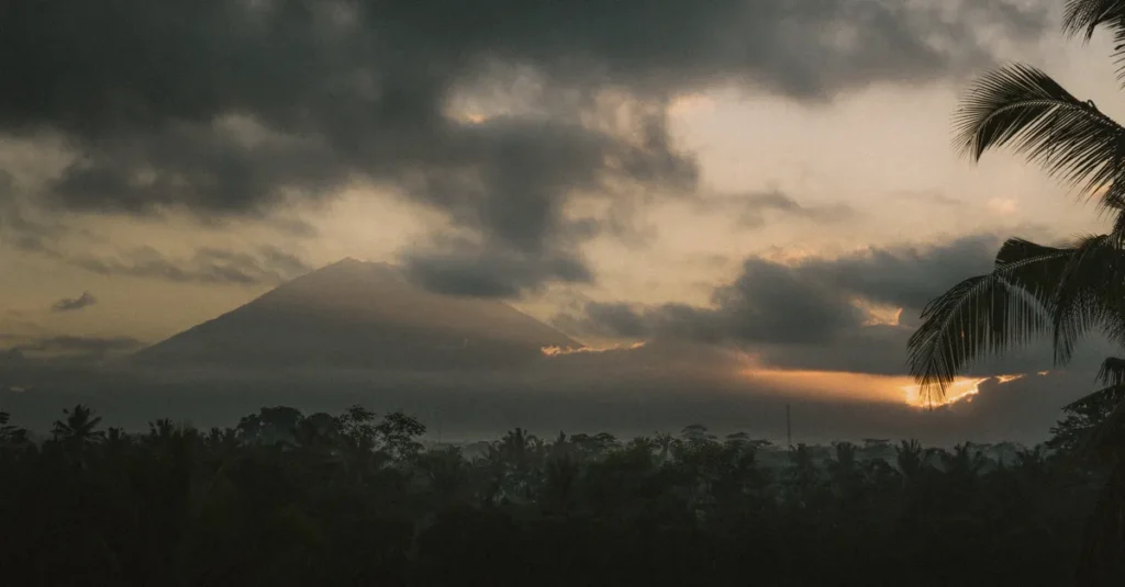 Indonésie en couple : volcans au lever, lagons au coucher (et Bali sans tomber dans les pièges) Witness the breathtaking sunrise over Mount Agung in Bali, enveloped by dramatic clouds and lush palms.