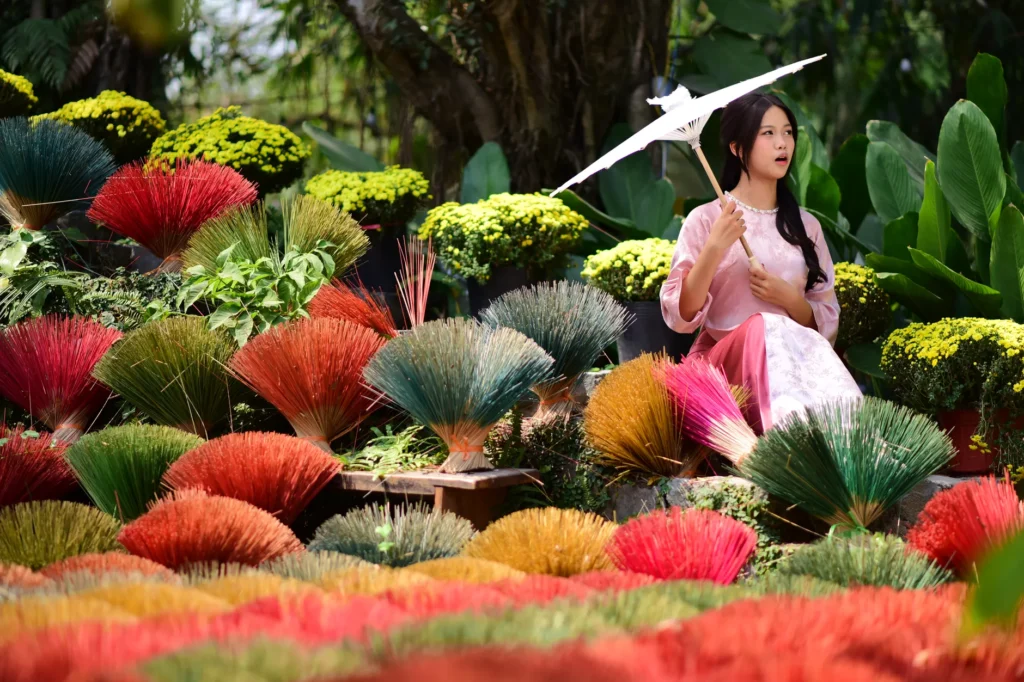 Woman in Vietnamese traditional attire sitting amidst colorful incense sticks, holding an umbrella.