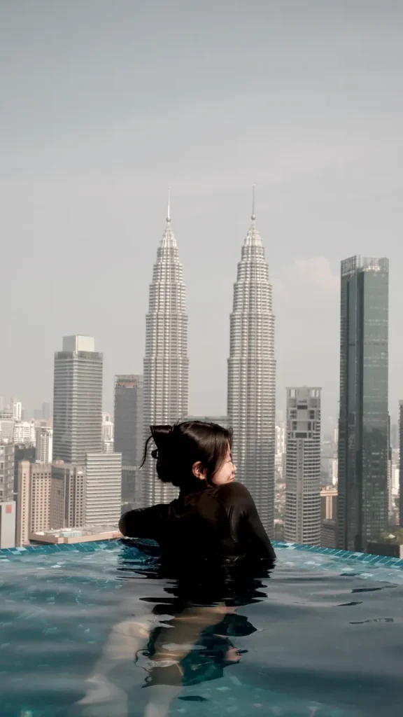 Woman relaxing in rooftop pool with view of Kuala Lumpur skyline including Petronas Towers.