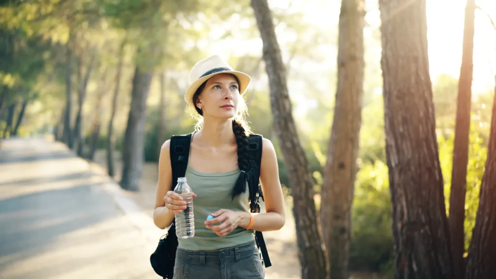Woman with braid and hat hiking in a forest, holding a water bottle during a sunlit evening.