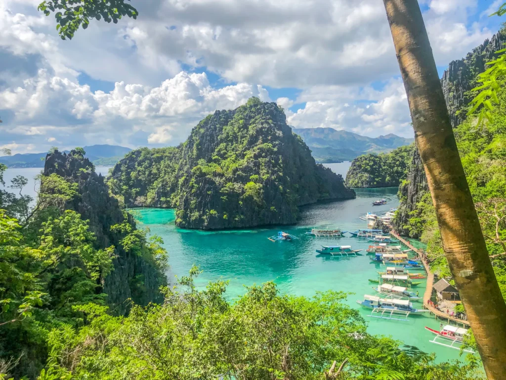 Explore a stunning tropical lagoon with turquoise waters, boats, and lush limestone cliffs under a bright cloudy sky.